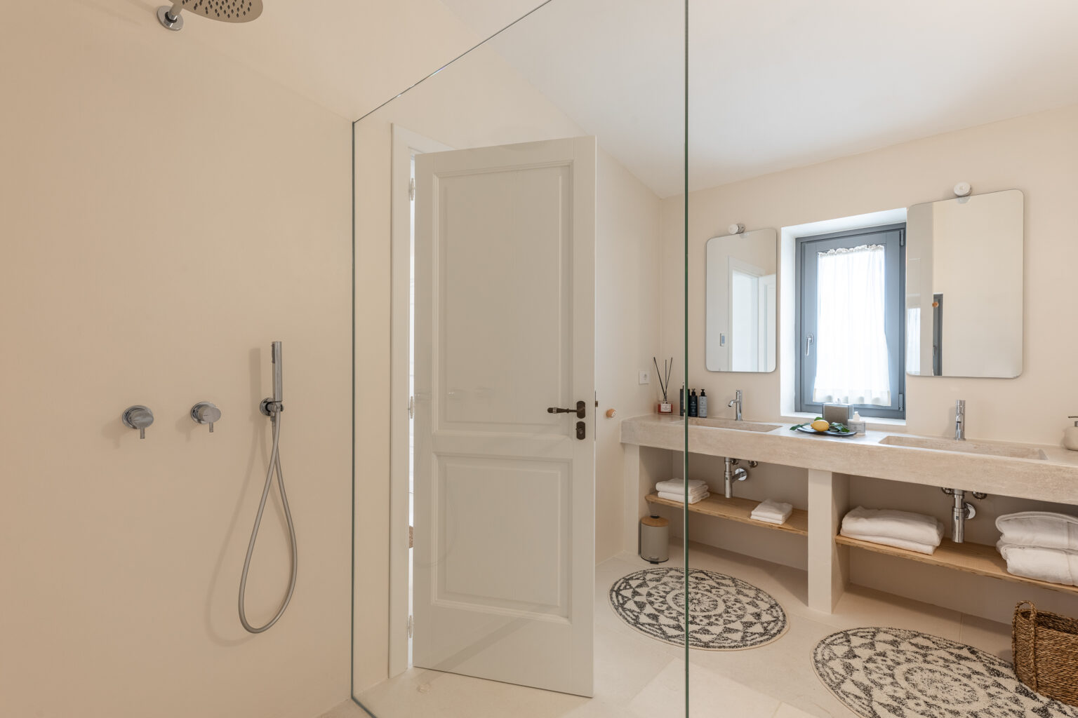 Modern bathroom interior featuring a glass shower enclosure, white cabinetry, and decorative rugs. Natural light streams through a window, highlighting the minimalist design and organized space with towels and toiletries. | Only on SopranoVillas