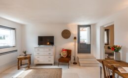 Cozy interior of a modern living space featuring a wooden dresser, flat-screen TV, armchair, and decorative items, with natural light streaming through the window and an inviting door leading outside.