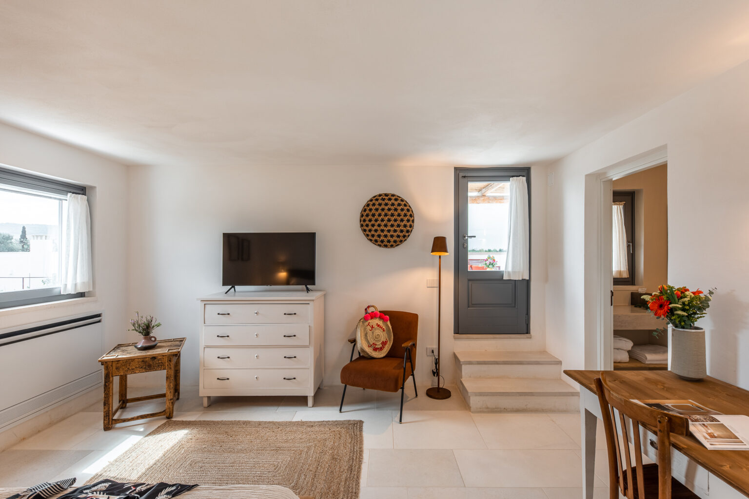 Cozy living room interior featuring a television, a wooden dresser, and a comfortable chair. Natural light fills the space, highlighting the minimalist decor and a small dining table with fresh flowers. The room has a welcoming atmosphere with neutral tones and stylish accents. | Only on SopranoVillas
