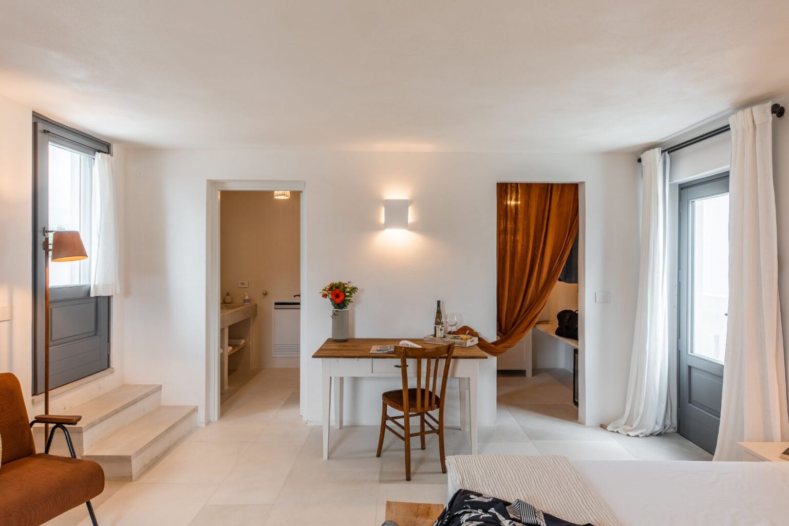 Bright and airy hotel room featuring a cozy seating area, a wooden desk with flowers, and large windows with sheer curtains. The space includes a stylish bathroom doorway and modern decor, ideal for a relaxing getaway. | Only on SopranoVillas