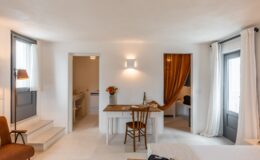 Bright and airy hotel room featuring a cozy seating area, a wooden desk with flowers, and large windows with sheer curtains. The space includes a stylish bathroom doorway and modern decor, ideal for a relaxing getaway.