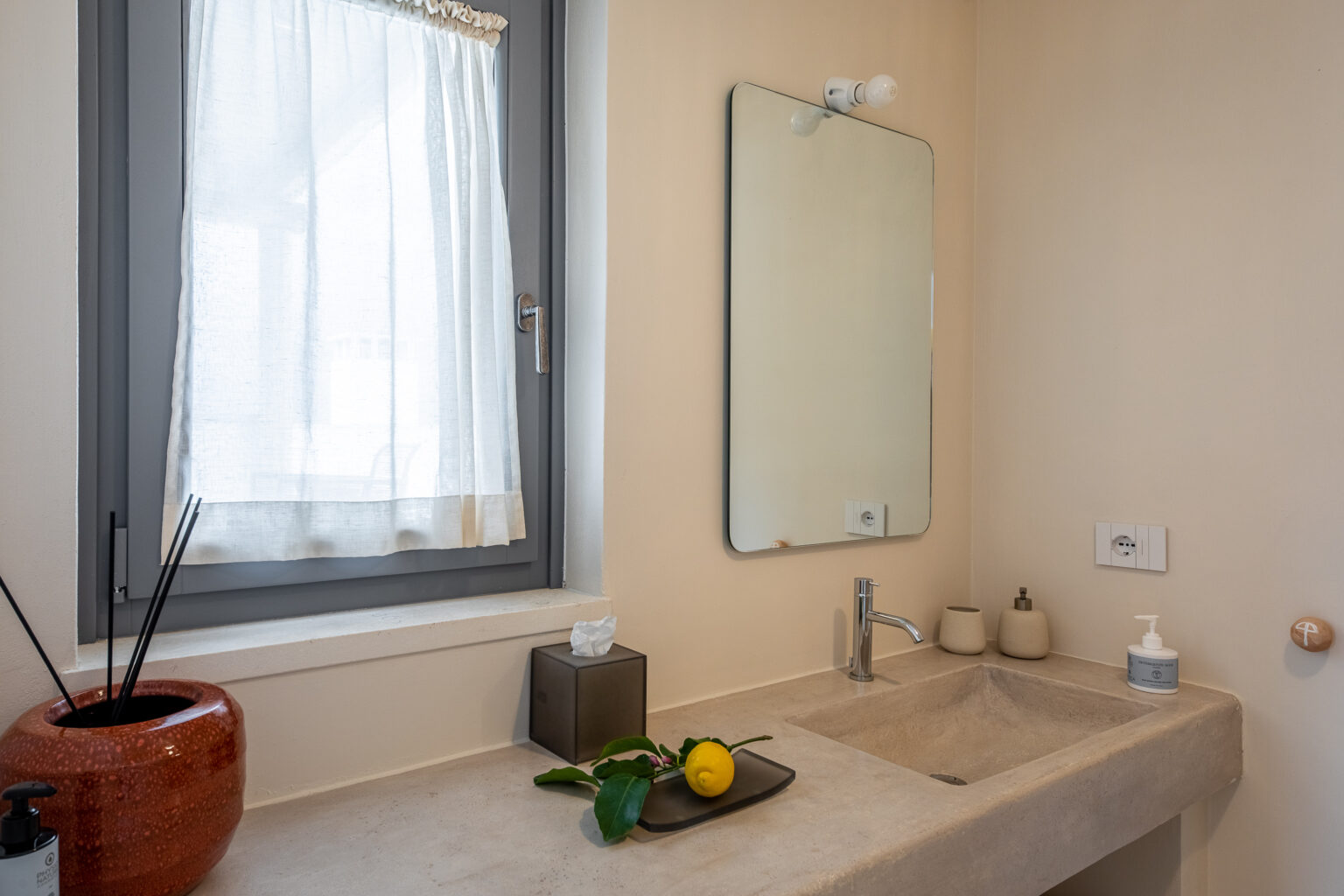 Modern bathroom interior featuring a sleek stone countertop, a large mirror, and natural light from a window with sheer curtains. The space is decorated with minimalistic accessories, including a decorative vase, a soap dispenser, and a small tray with a lemon and green leaves, creating a fresh and inviting atmosphere. | Only on SopranoVillas