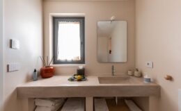 Modern bathroom design featuring a sleek stone sink, a large mirror, and natural light from a window. The space includes neatly arranged towels, aromatic elements like a decorative vase, and personal care products, creating a clean and inviting atmosphere.