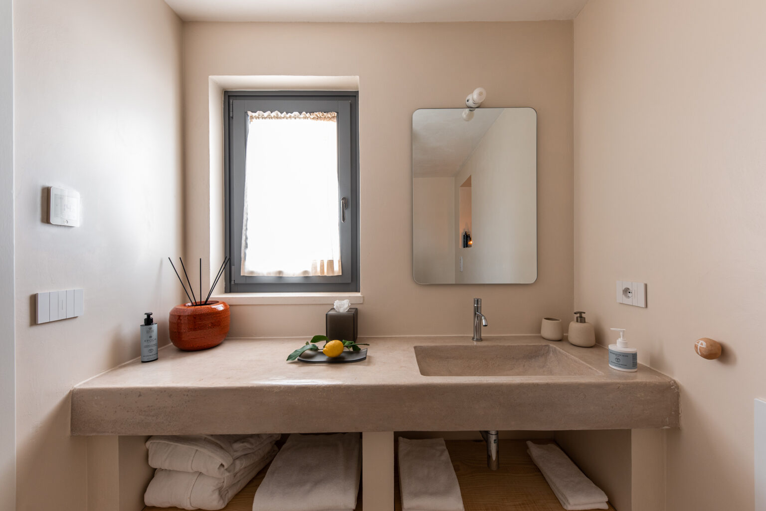 Modern bathroom with a minimalist design featuring a concrete countertop, a large mirror, and natural light from a window. The space includes decorative items like a fruit bowl and candles, with neatly arranged towels stored below. | Only on SopranoVillas