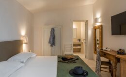 Cozy hotel room featuring a double bed with white linens, a green textured runner, and decorative items including a hat and sunglasses. The room includes a wooden desk, a mirror, and access to a modern bathroom visible through an open door. Soft lighting enhances the relaxing atmosphere.