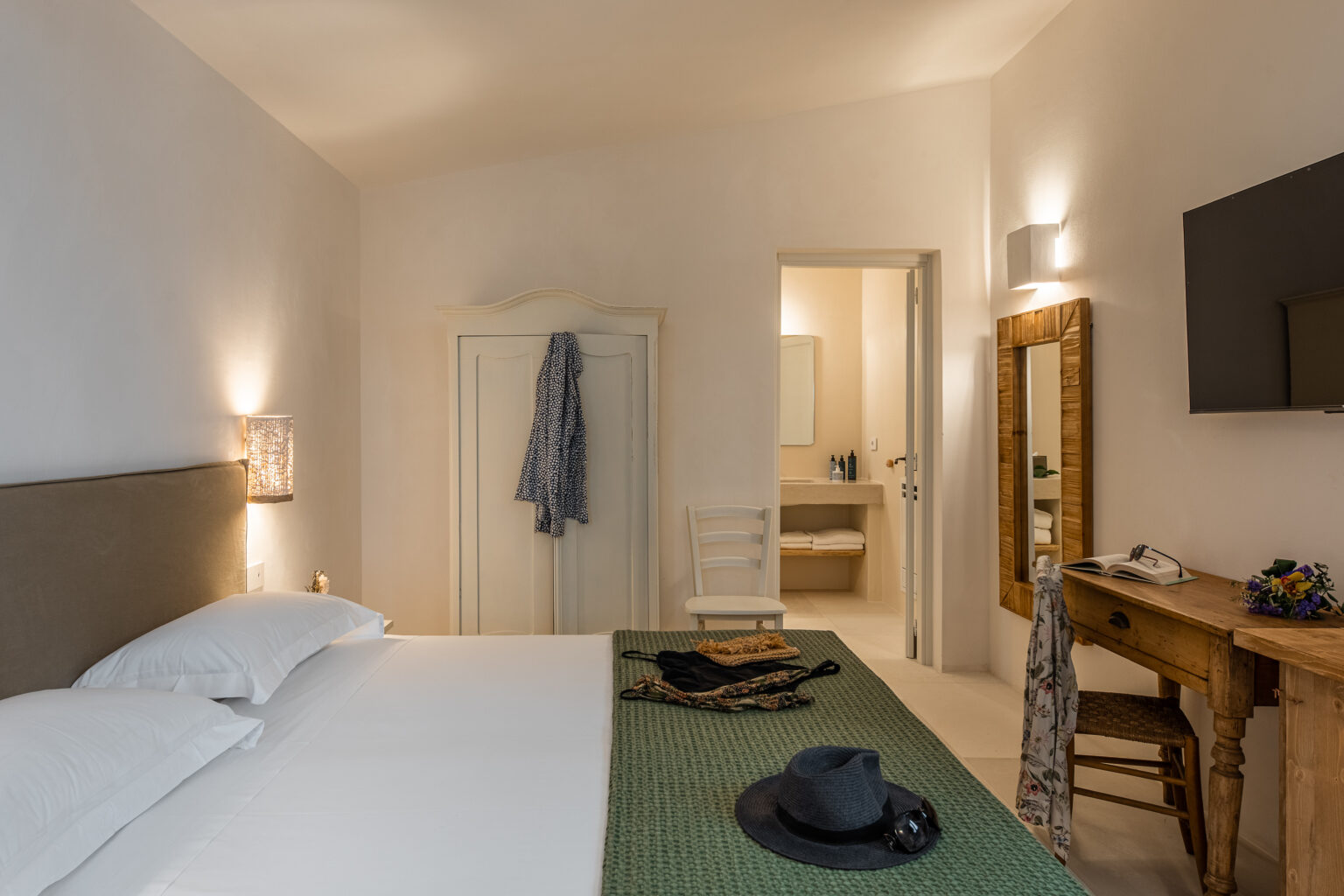 Cozy hotel room featuring a comfortable bed with white linens, a green accent blanket, a wooden desk with a chair, and a flat-screen TV. The room includes a doorway leading to a bathroom and is decorated with minimalistic furnishings, creating a serene atmosphere. | Only on SopranoVillas