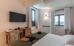 Cozy bedroom interior featuring a large window with white curtains, a wooden side table, a wall-mounted TV, and a neatly made bed with green and white bedding. Decorative elements include sunglasses, a hat, and accessories on the bed, creating a welcoming atmosphere. Natural light enhances the bright and airy feel of the space.