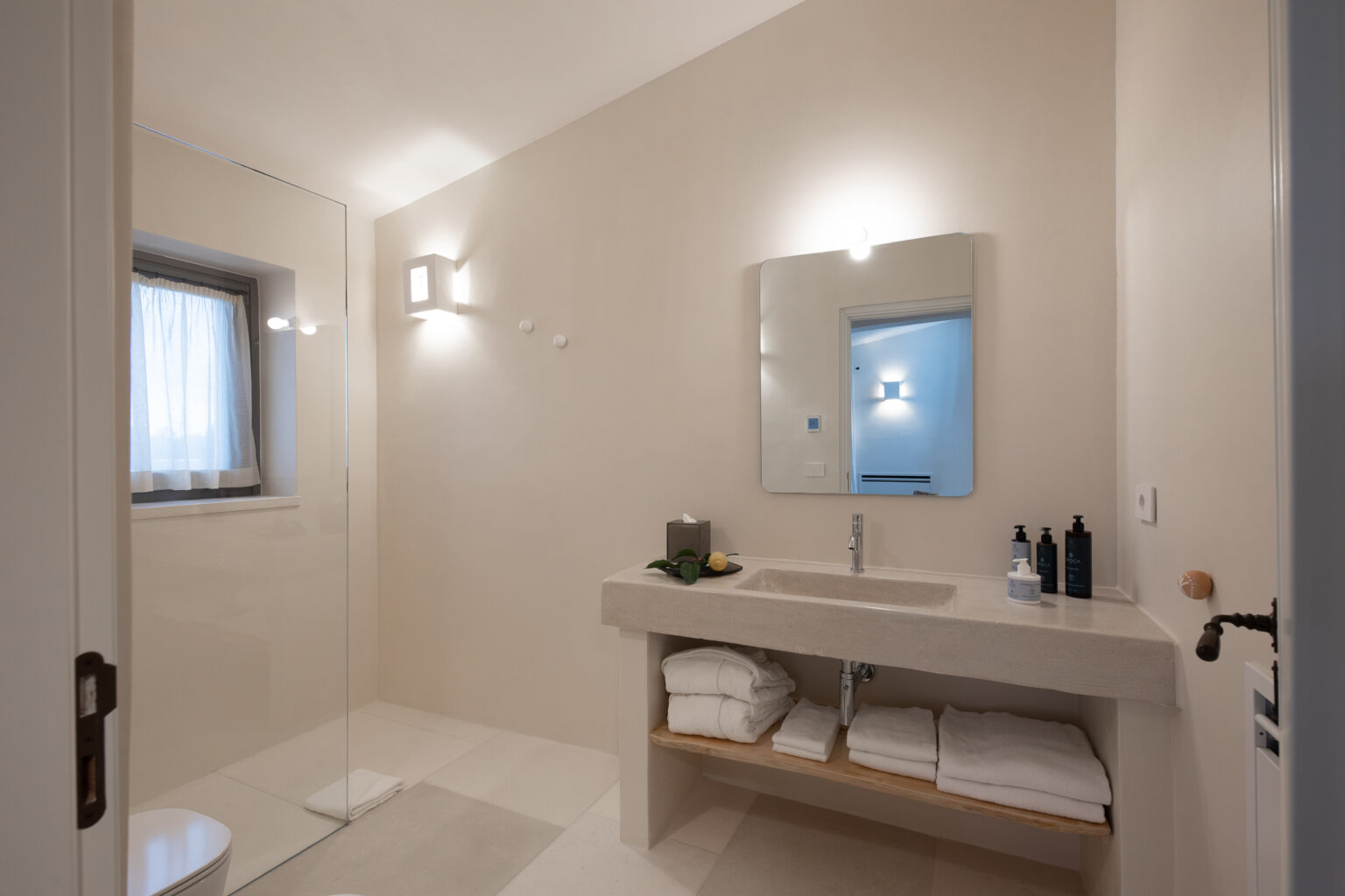 Modern bathroom interior featuring a glass shower, minimalist design, and natural light from a window. The stone sink is accompanied by neatly arranged towels and skincare products on a wooden shelf. Soft lighting enhances the serene atmosphere. | Only on SopranoVillas