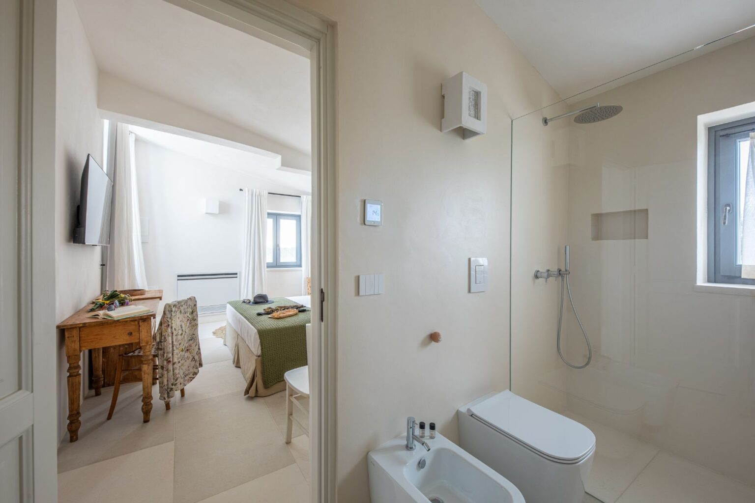 Modern bathroom with a glass shower and bidet, leading into a bright, airy bedroom featuring a wooden desk, floral chair, and a cozy bed with green bedding, ideal for travelers seeking comfort and style. | Only on SopranoVillas