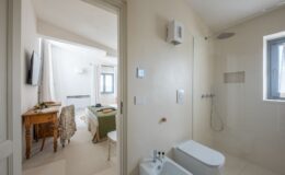 Modern bathroom with a glass shower and bidet, leading into a bright, airy bedroom featuring a wooden desk, floral chair, and a cozy bed with green bedding, ideal for travelers seeking comfort and style.