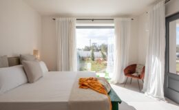Bright and airy bedroom featuring a king-size bed with decorative pillows, a green bench adorned with a cozy throw, and a stylish brown chair. Large windows with sheer curtains offer a view of a sunny garden with olive trees and lounge chairs, creating a serene and inviting atmosphere.