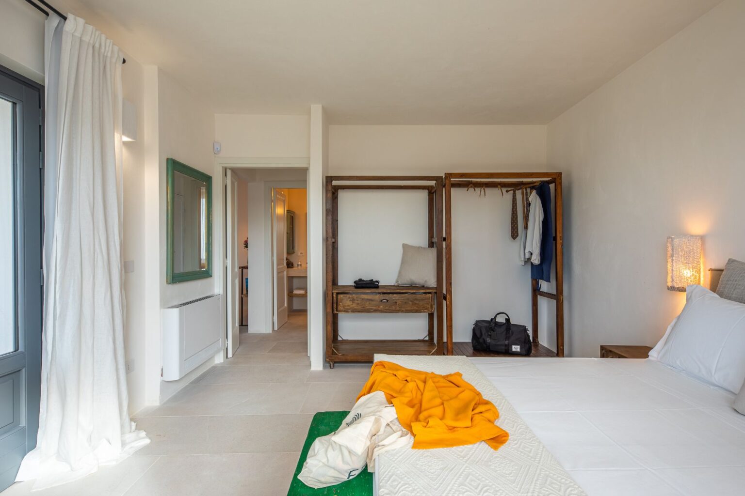 Modern bedroom interior featuring a bed with white linens, an orange throw, and wooden furniture. Open wardrobe displays clothes and accessories, while a large window allows natural light. The minimalist decor includes a green-framed mirror and a cozy atmosphere. Suitable for showcasing stylish and functional bedroom design ideas. | Only on SopranoVillas