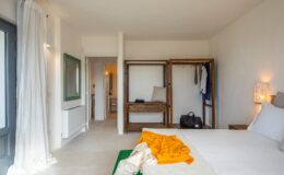 Modern bedroom interior featuring a bed with white linens, an orange throw, and wooden furniture. Open wardrobe displays clothes and accessories, while a large window allows natural light. The minimalist decor includes a green-framed mirror and a cozy atmosphere. Suitable for showcasing stylish and functional bedroom design ideas.