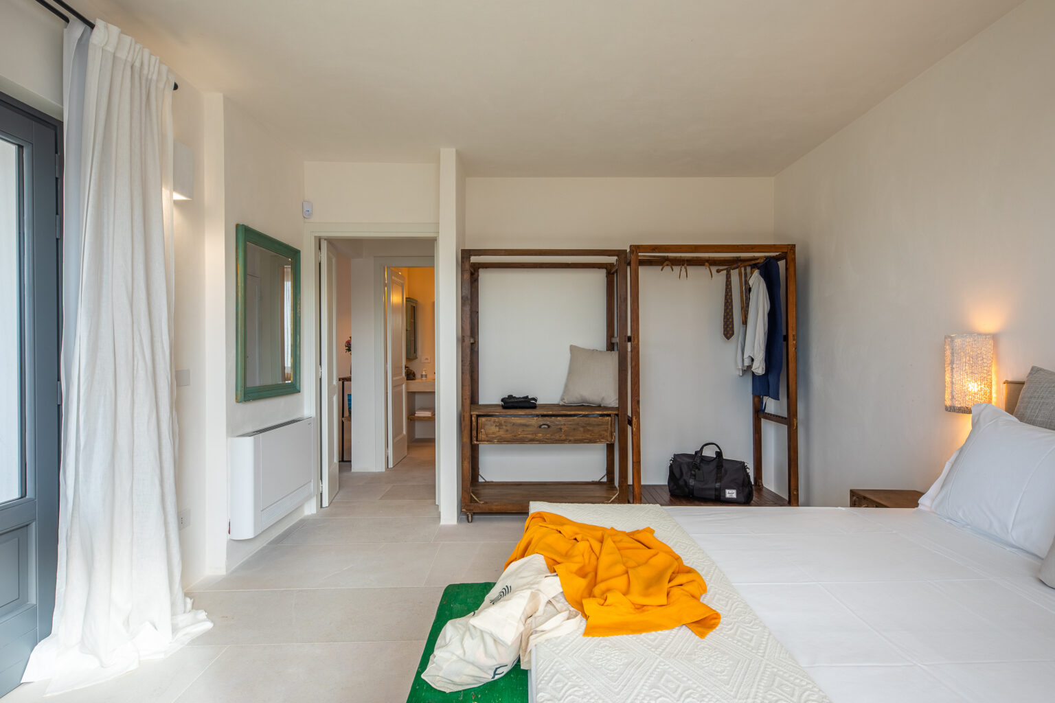 Modern hotel room featuring a spacious layout, minimalist decor, a comfortable bed with orange and green accents, and wooden clothing racks. Natural light filters through sheer curtains, creating a bright and inviting atmosphere. | Only on SopranoVillas