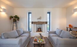 Cozy living room featuring two light gray sofas, a rustic coffee table with fruit and snacks, and decorative elements like a wooden shelf and potted plants. Natural light filters through the windows, enhancing the inviting atmosphere.