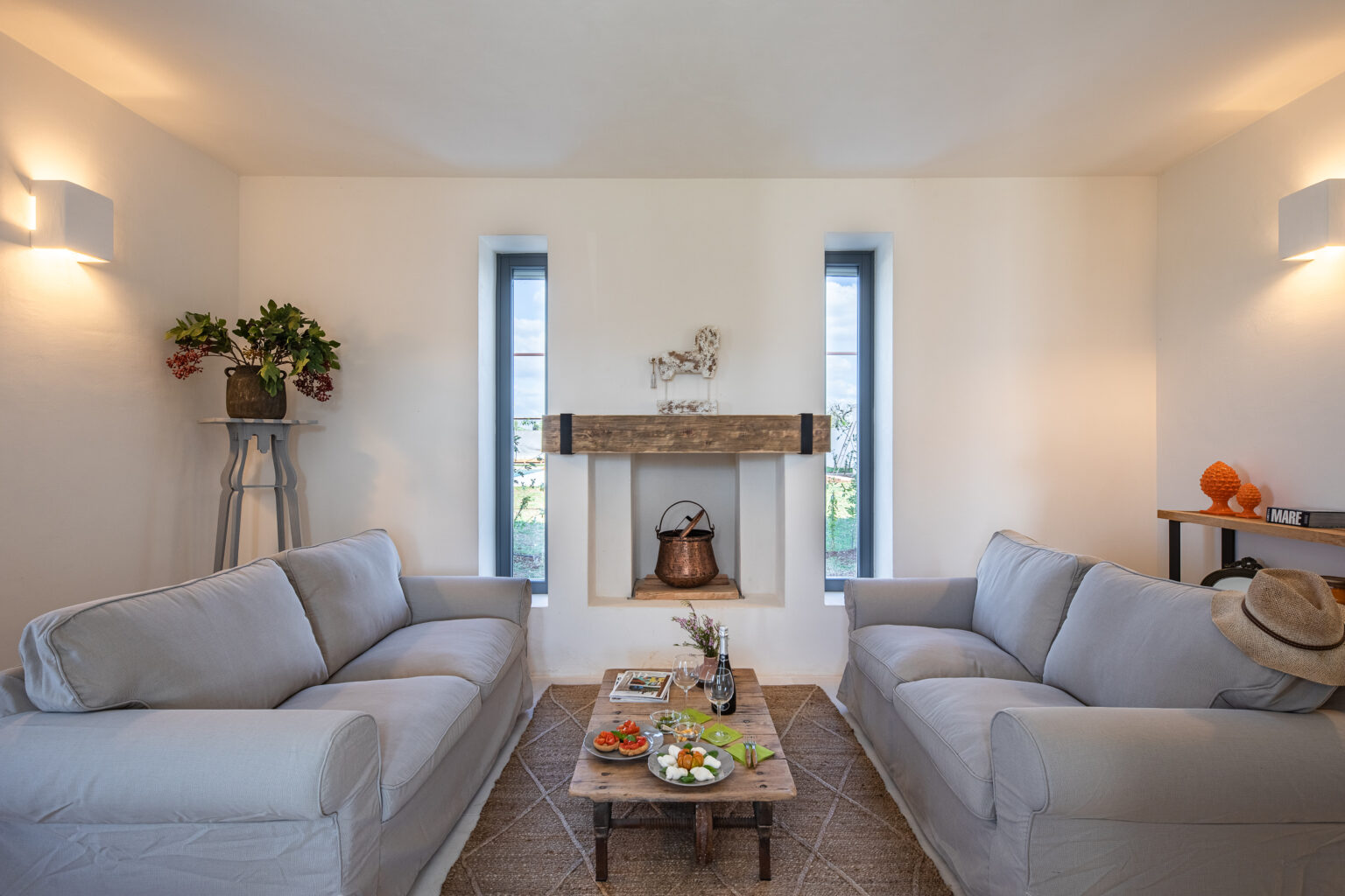 Cozy living room featuring two light gray sofas, a wooden coffee table with snacks, and a decorative fireplace. Natural light streams through the windows, highlighting a minimalist design with plants and unique decor. Ideal for relaxation and social gatherings. | Only on SopranoVillas
