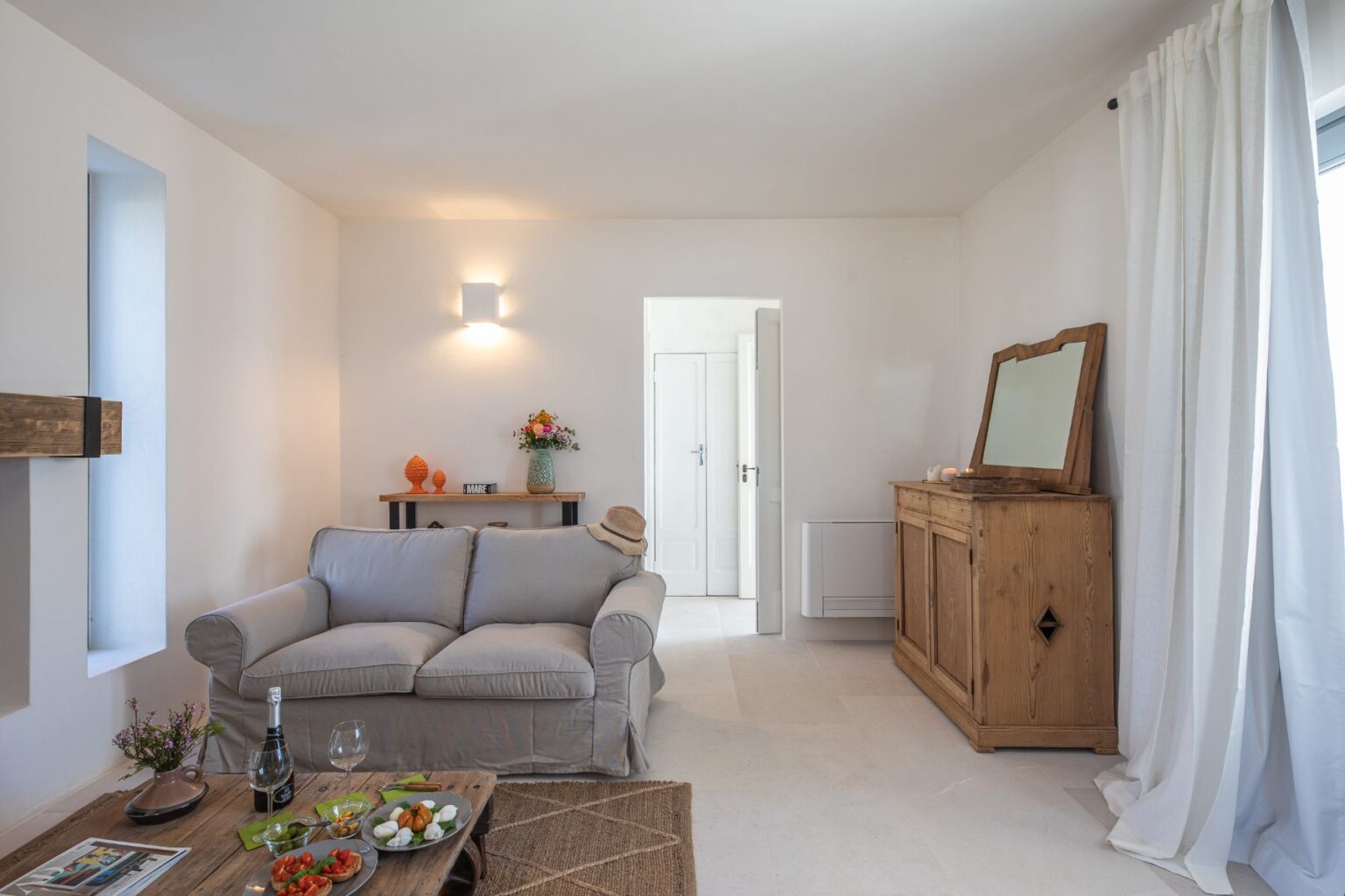 Bright and airy living room featuring a gray sofa, wooden coffee table with snacks, and floral decor. The room also includes a wooden sideboard, a hat on the sofa, and natural light flowing in through a window, creating a cozy and inviting atmosphere. | Only on SopranoVillas