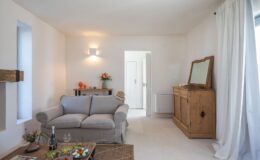 Bright and airy living room featuring a gray sofa, wooden coffee table with snacks, and floral decor. The room also includes a wooden sideboard, a hat on the sofa, and natural light flowing in through a window, creating a cozy and inviting atmosphere.