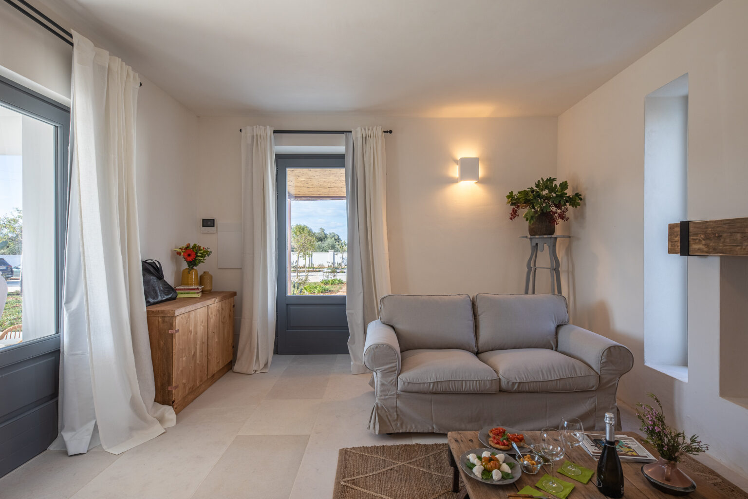 Cozy living room featuring a light gray sofa, wooden sideboard, and a rustic coffee table set with snacks and drinks. Large windows and white curtains allow natural light, enhancing the inviting atmosphere. A potted plant and soft lighting add to the decor. | Only on SopranoVillas