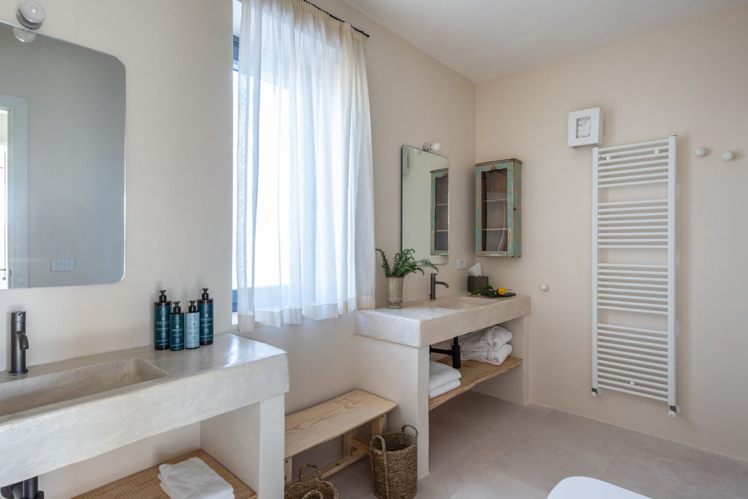 Modern bathroom interior featuring a minimalist design with a concrete sink, wooden shelving, and a large mirror. Natural light filters through a sheer curtain, illuminating toiletries and fresh greenery on the counter. The space is enhanced with a white towel rack and rustic wooden accents, creating a serene atmosphere. | Only on SopranoVillas