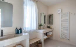 Modern bathroom interior featuring a minimalist design with a concrete sink, wooden shelving, and a large mirror. Natural light filters through a sheer curtain, illuminating toiletries and fresh greenery on the counter. The space is enhanced with a white towel rack and rustic wooden accents, creating a serene atmosphere.