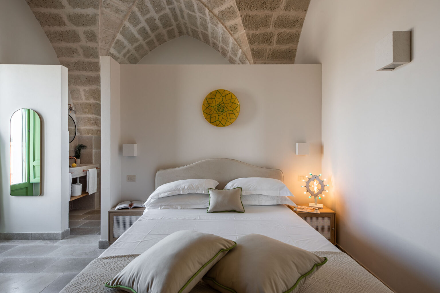 Cozy bedroom featuring a plush bed with white linens and decorative pillows, stone arch ceiling, and a vibrant yellow wall decoration. The room includes a modern mirror and green window, with a bedside table adorned with a stylish lamp and a book, creating a serene and inviting atmosphere. | Only on SopranoVillas