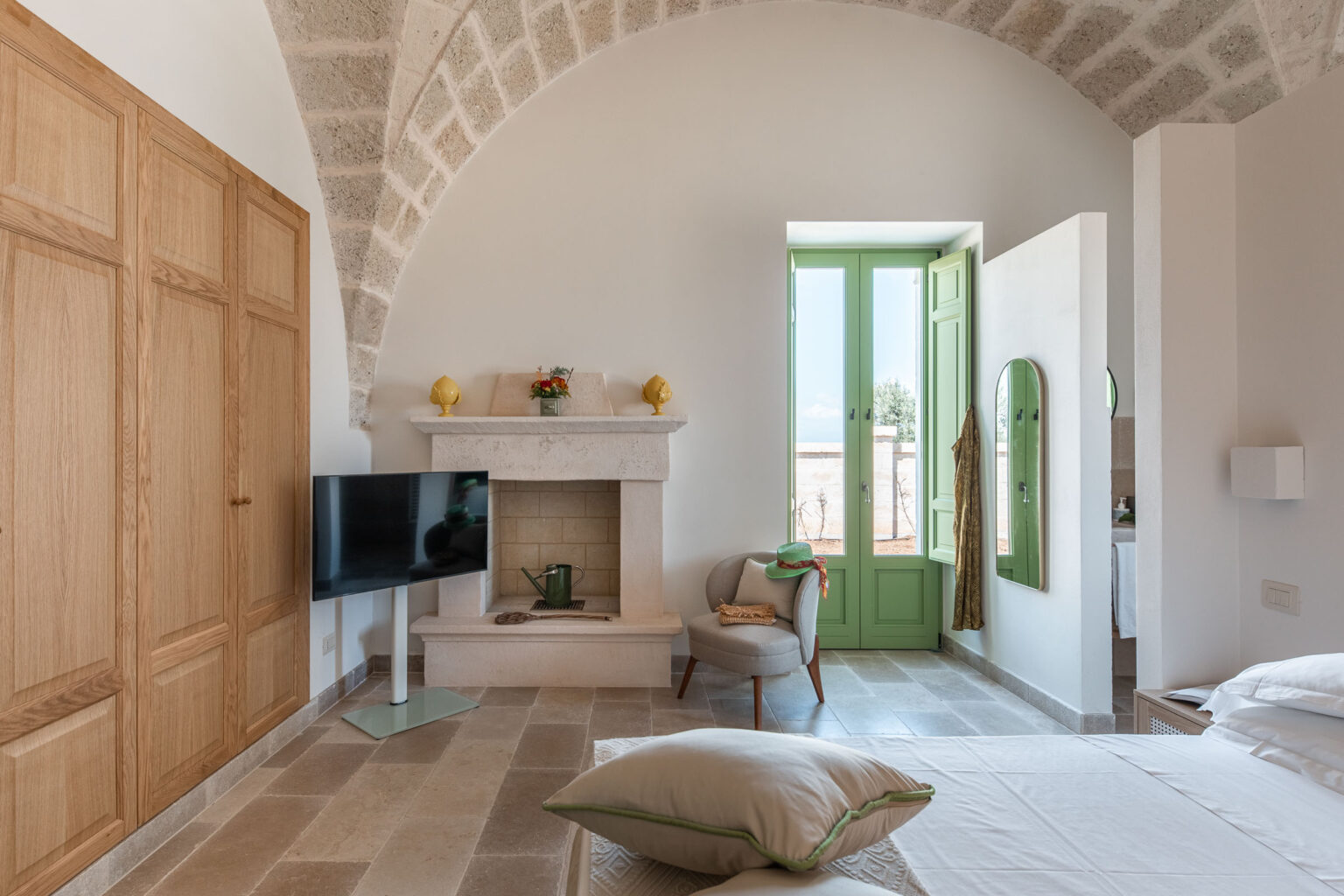 Bright and airy bedroom featuring a stone fireplace, wooden wardrobe, and a green window that opens to an outdoor view. The room includes a comfortable chair, a modern TV, and decorative elements that enhance its cozy atmosphere. | Only on SopranoVillas