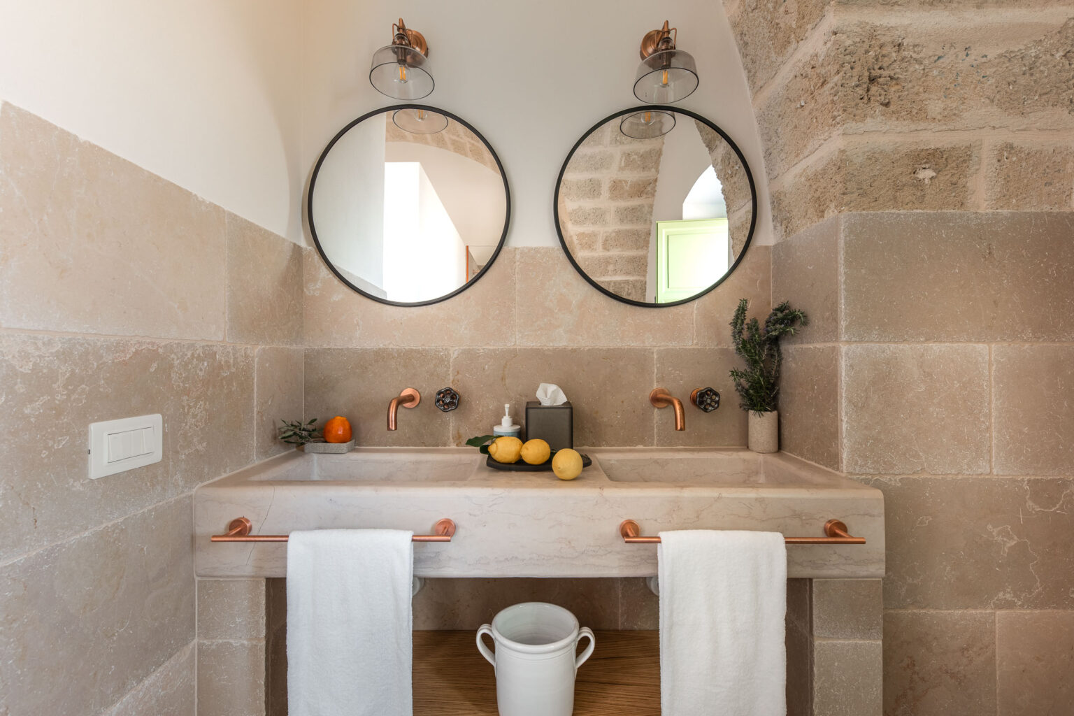 Modern bathroom design featuring a double sink with a marble countertop, copper faucets, and stylish round mirrors. The space includes natural elements like lemons and an orange, complemented by white towels and a decorative plant. The warm stone walls and wooden accents enhance the contemporary aesthetic. | Only on SopranoVillas