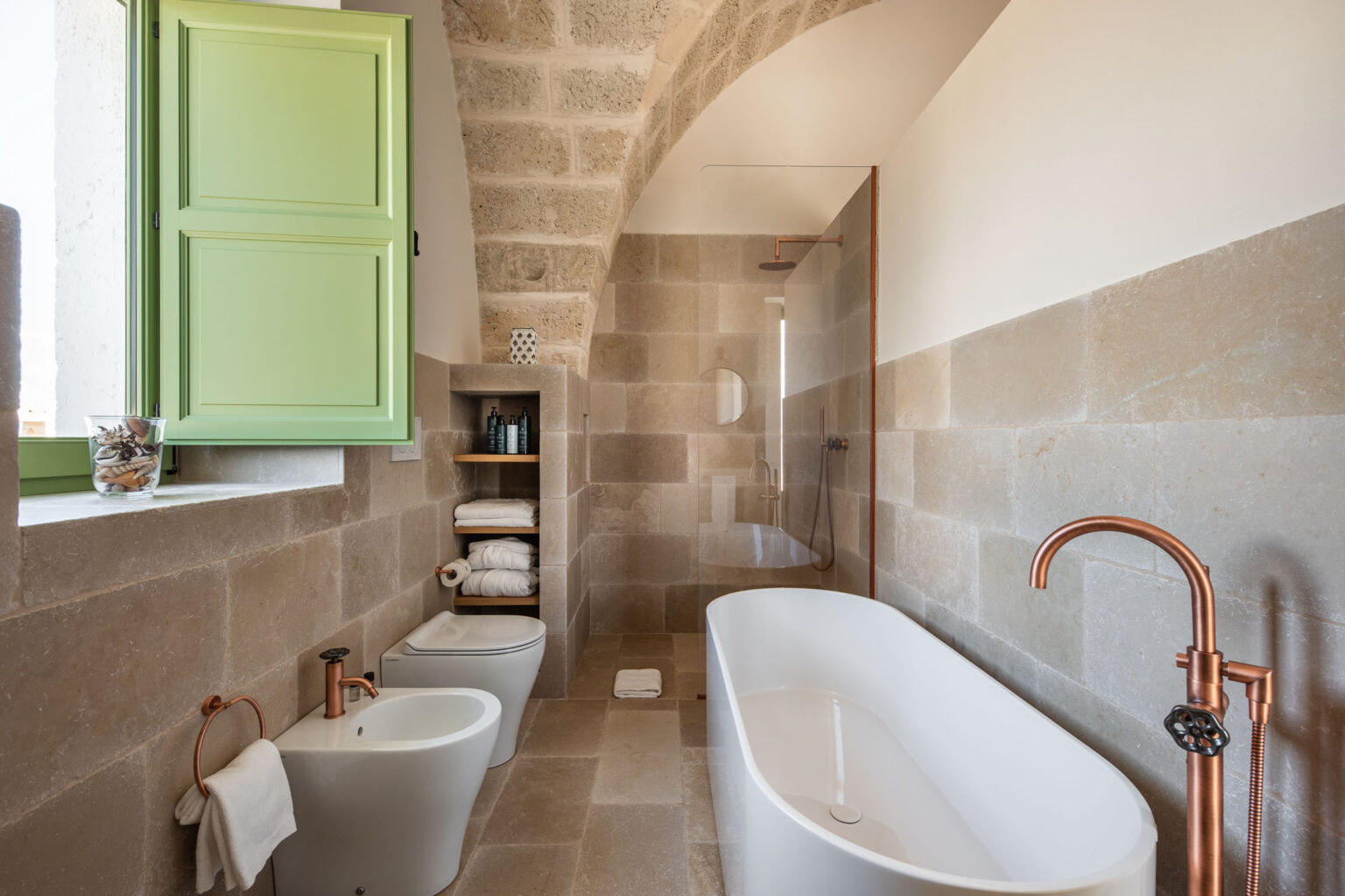 Modern bathroom featuring a freestanding white bathtub, a stylish copper faucet, and a bidet. The space includes natural stone walls, neatly stacked towels, and a green window, providing a bright and airy ambiance. | Only on SopranoVillas