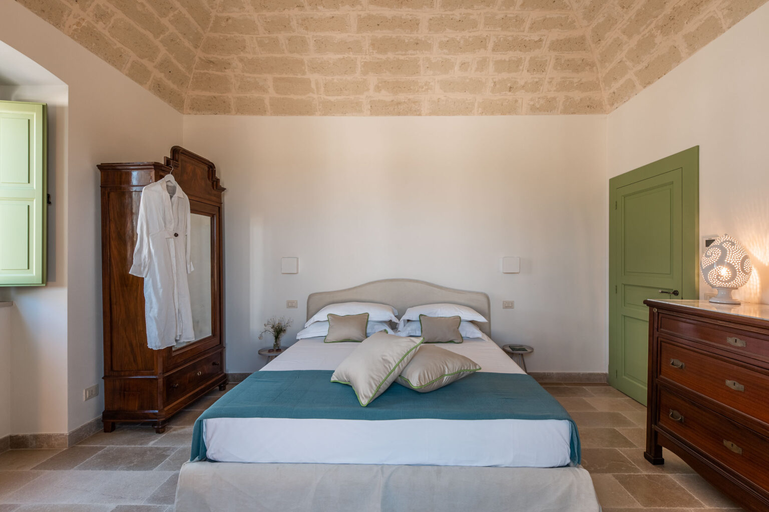 Cozy bedroom featuring a comfortable bed with decorative pillows, a vintage wooden wardrobe, and a stylish lamp. The room has a high vaulted ceiling and light-colored stone walls, complemented by green accents in the window and door. Ideal for relaxation and comfort. | Only on SopranoVillas