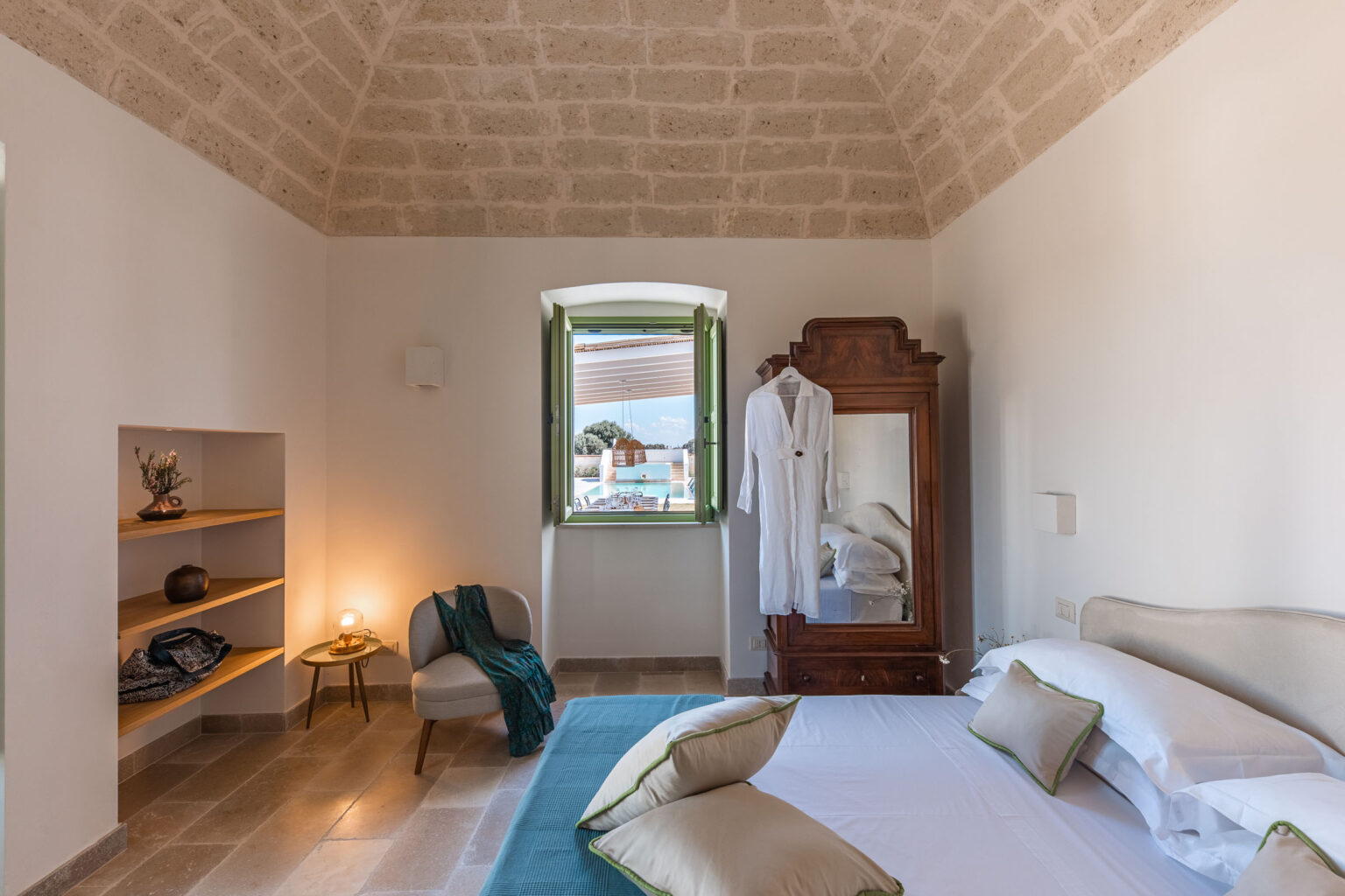 Bright and airy bedroom featuring a high vaulted stone ceiling, a cozy bed with decorative pillows, a vintage wardrobe, and a window with green shutters offering a view of the outdoors. A small reading chair and wooden shelves add to the inviting atmosphere. | Only on SopranoVillas