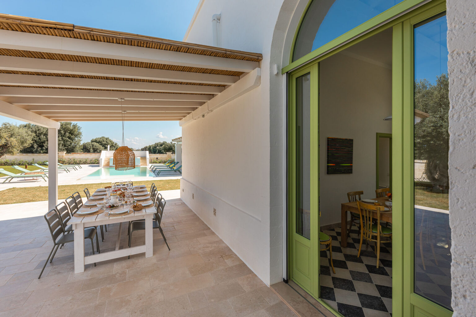 Outdoor dining area with a large table set for a meal, featuring a pool and lounge chairs in the background. The scene includes a modern architectural design with a vibrant green door leading to an interior dining space. | Only on SopranoVillas