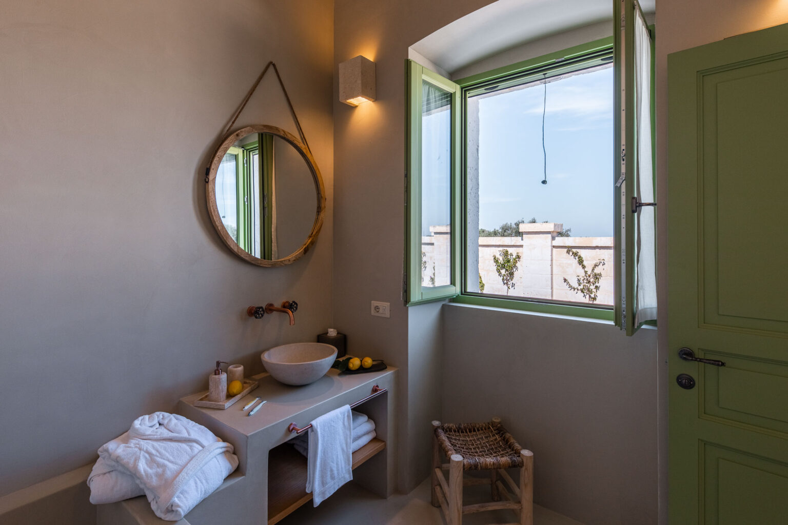 Modern bathroom interior featuring a round wooden mirror, a stylish sink, and green-framed windows. Soft towels and fresh lemons add a touch of elegance, while natural light enhances the serene atmosphere. | Only on SopranoVillas
