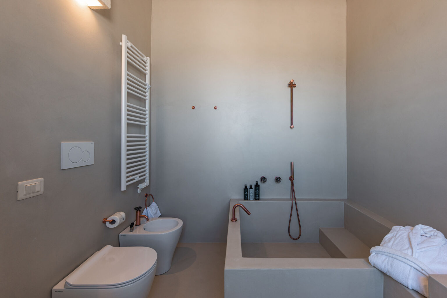 Modern bathroom design featuring a minimalist aesthetic with a concrete tub, copper fixtures, and a heated towel rack. The space includes a toilet and bidet, complemented by neatly arranged toiletries and a plush white towel. | Only on SopranoVillas