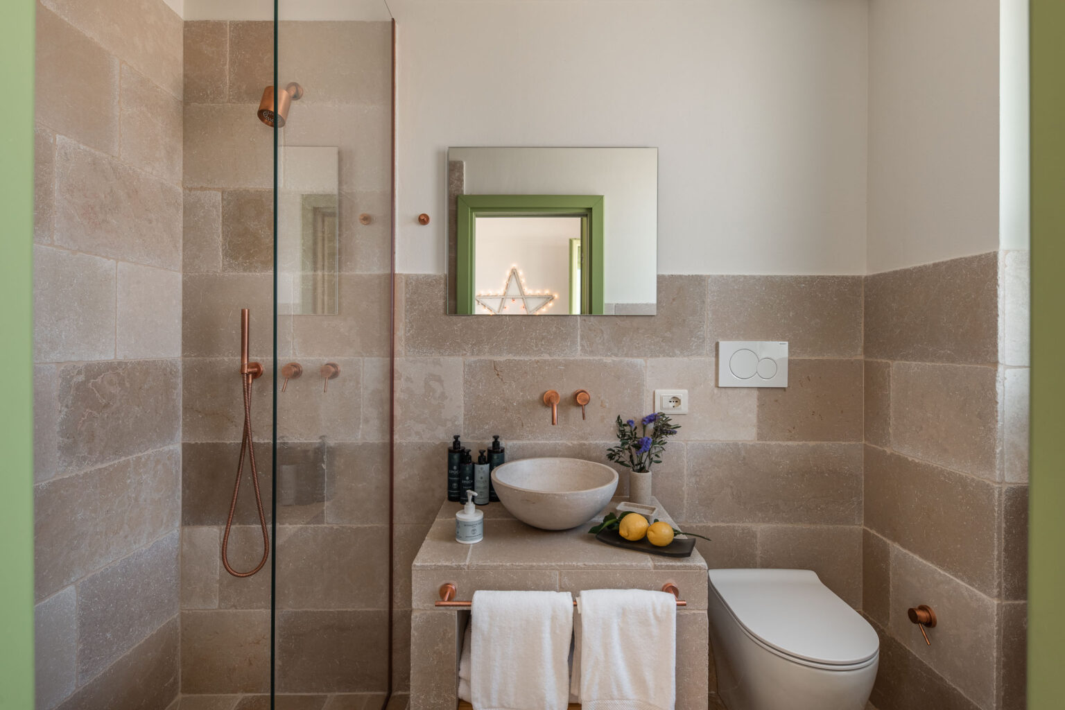 Modern bathroom featuring a glass shower with copper fixtures, a stylish stone countertop with a round basin, and decorative elements including a mirror with a green frame and fresh flowers. The space is accented by neutral stone tiles and a minimalist design, creating a serene atmosphere. | Only on SopranoVillas