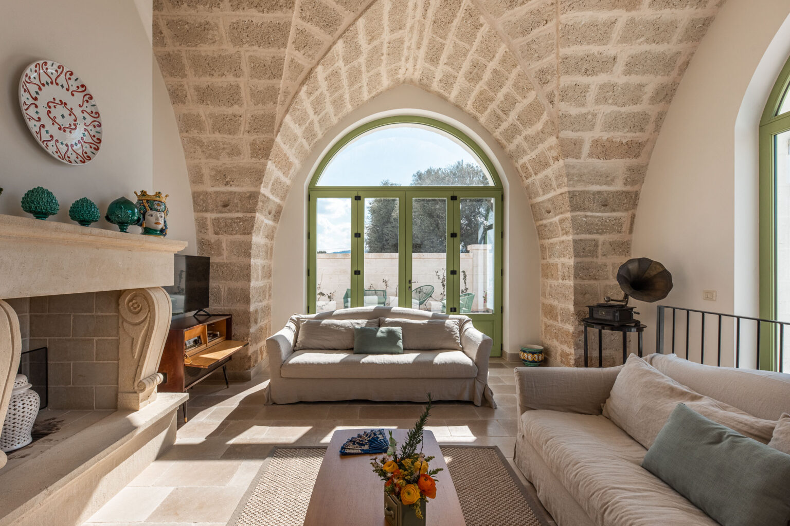Bright and airy living room featuring stone walls, large arched windows, and comfortable sofas. The interior design includes a modern coffee table, decorative accents, and natural light illuminating the space. | Only on SopranoVillas