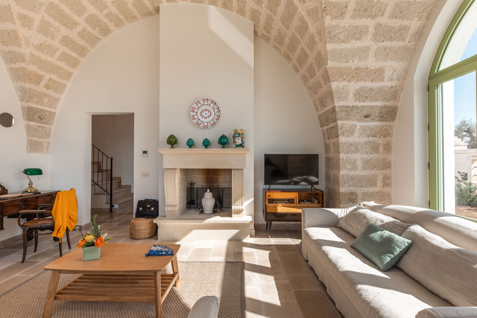 Bright and airy living room featuring a stone archway, a comfortable gray sofa, a wooden coffee table with decorative fruit, and a modern television. The space is adorned with a vibrant wall clock and green accents, creating a cozy and inviting atmosphere. | Only on SopranoVillas
