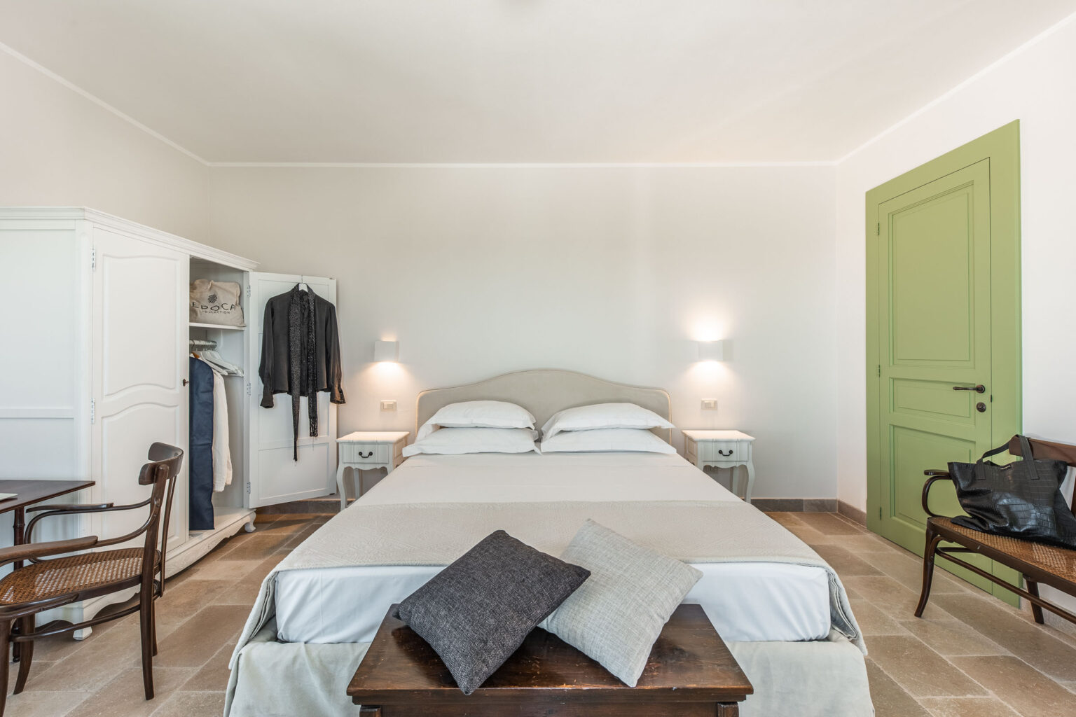 Modern bedroom interior featuring a double bed with white linens, two bedside tables with lamps, a wooden table and chair, and a stylish wardrobe. The room has a soft color palette, natural light, and a green door, creating a serene and inviting atmosphere. | Only on SopranoVillas