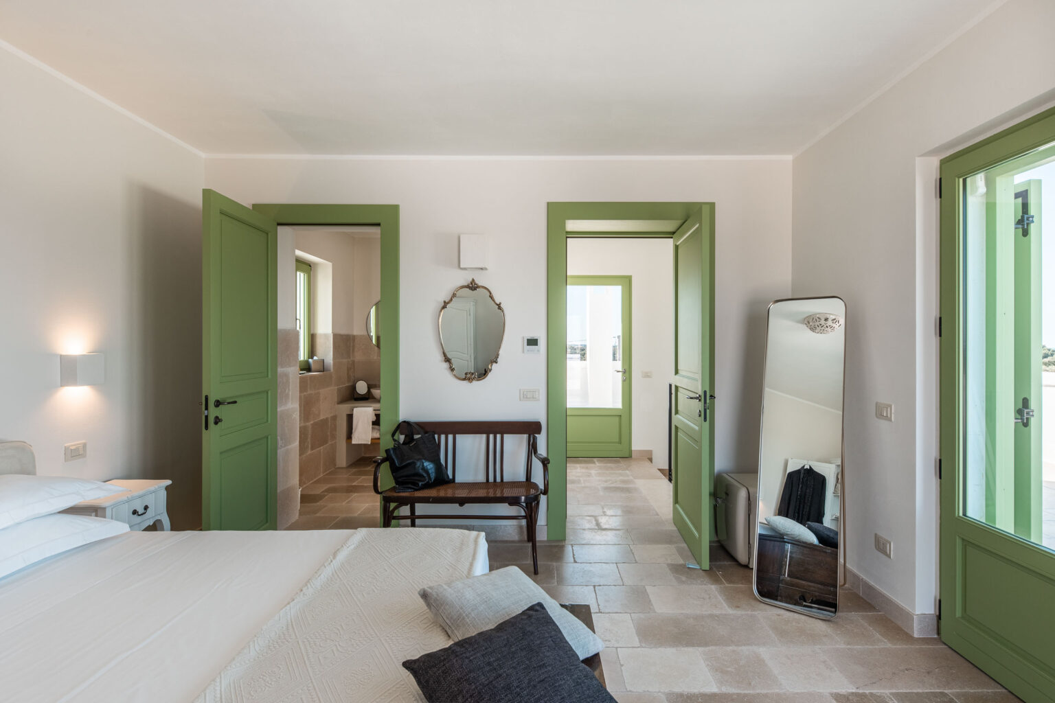 Bright and airy bedroom featuring green doors, a wooden bench, and a full-length mirror, with a view of a modern bathroom through an open doorway. The decor combines natural stone flooring and minimalist furnishings, creating a serene atmosphere. | Only on SopranoVillas