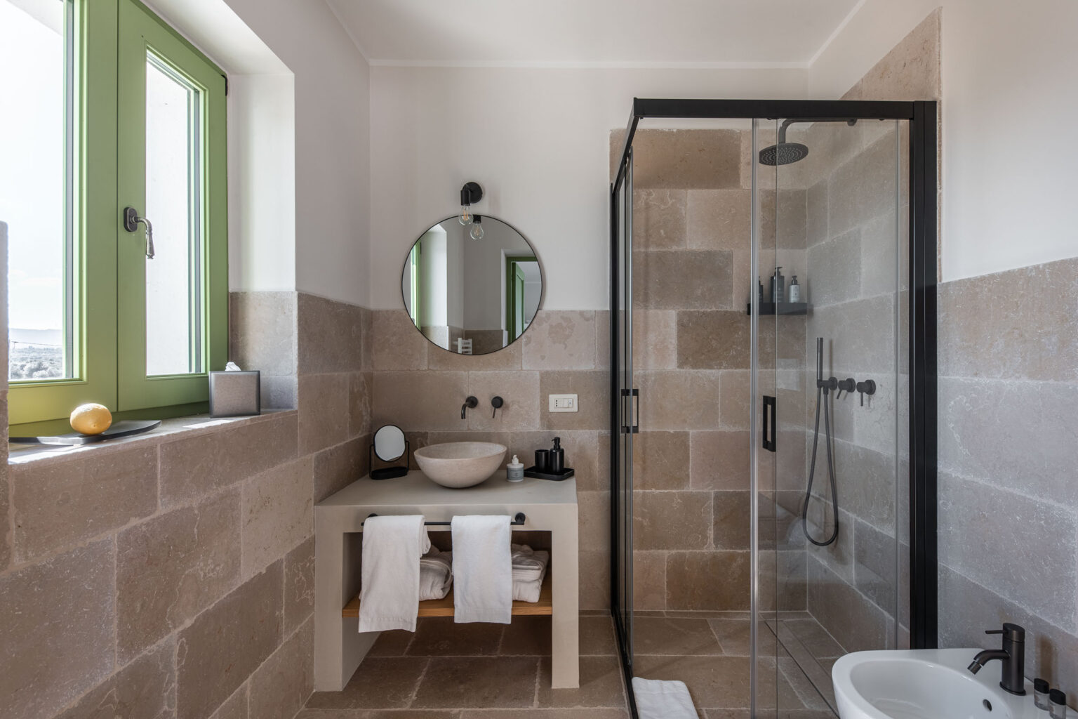 Modern bathroom featuring a glass shower enclosure, natural stone walls, a round mirror, and a stylish sink with wooden storage underneath. Bright green window frames enhance the natural light and aesthetic. | Only on SopranoVillas