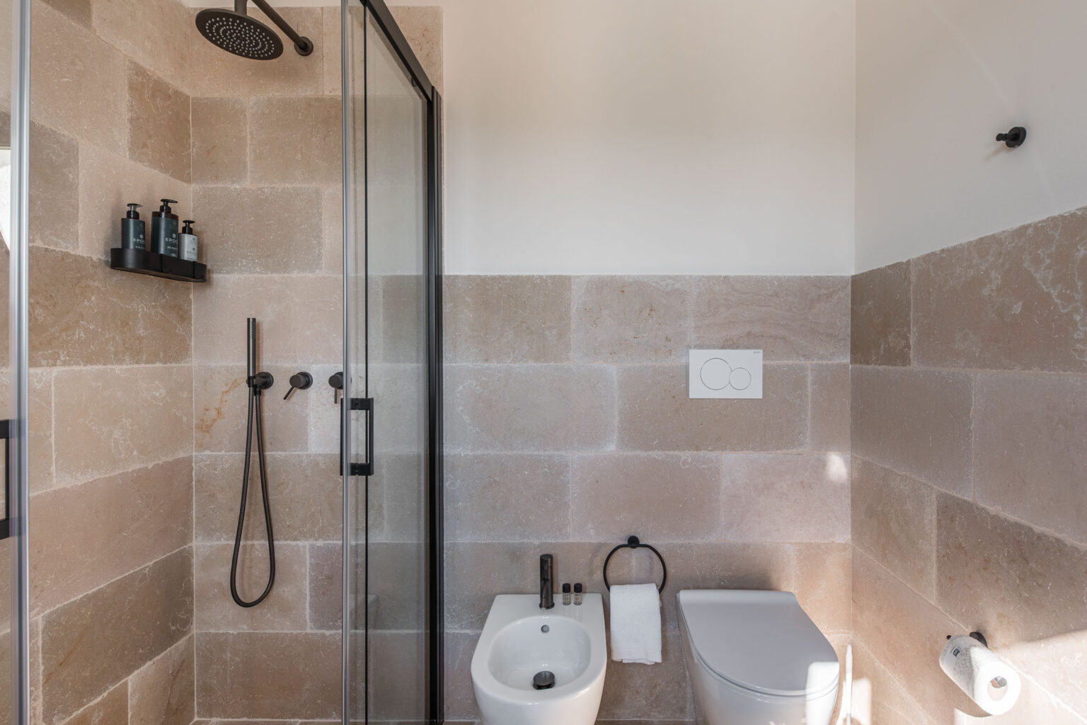 Modern bathroom interior featuring a glass shower enclosure, stone wall tiles, a wall-mounted shelf with bathroom products, a sleek toilet, and a towel holder. The design emphasizes a minimalist aesthetic with neutral tones and contemporary fixtures. | Only on SopranoVillas