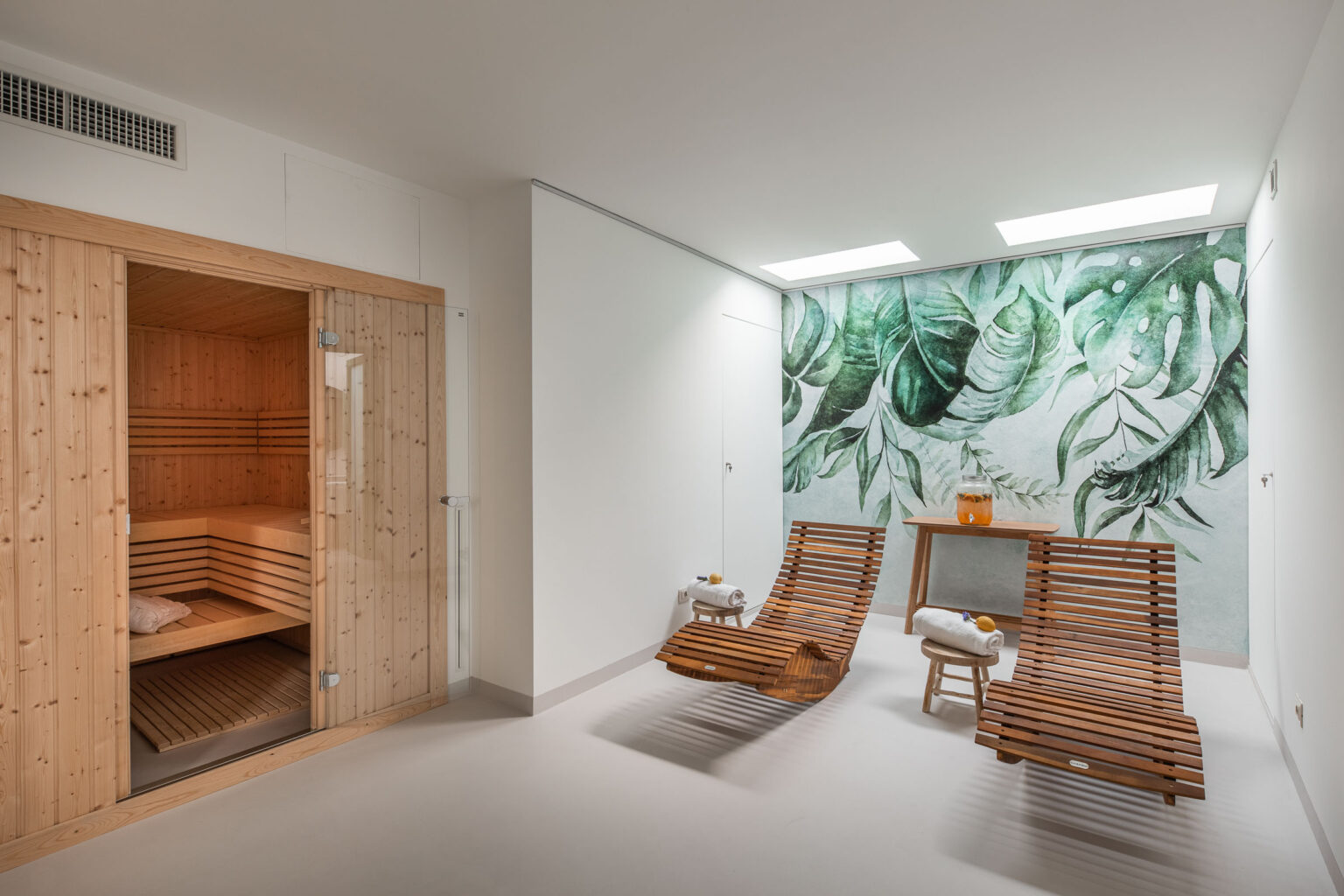 Modern wellness spa interior featuring a wooden sauna, two slatted lounge chairs, and a vibrant leaf mural. The space is designed for relaxation, with natural light streaming in and a calming atmosphere. | Only on SopranoVillas