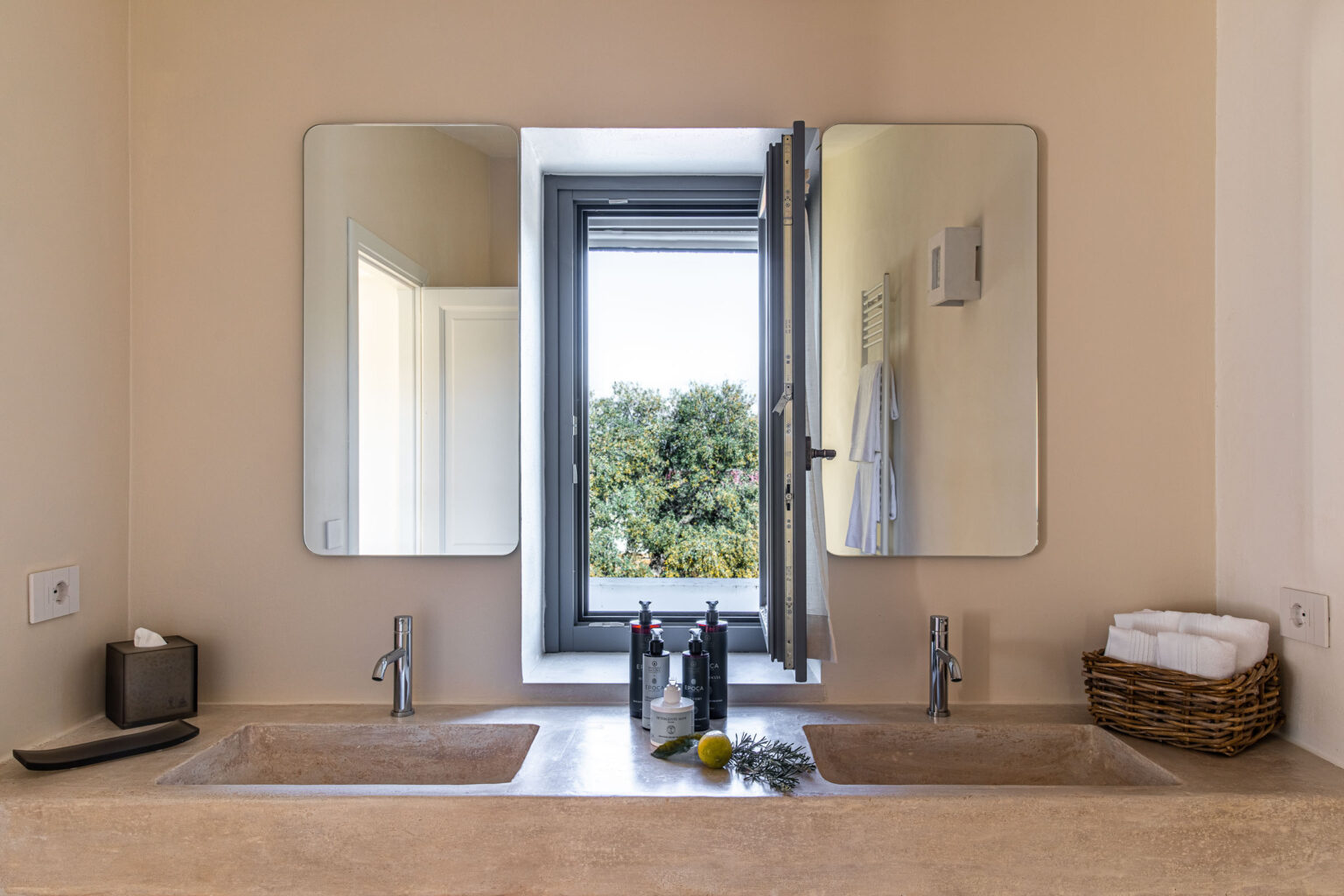 Modern bathroom with double sinks, large mirrors, and a window overlooking greenery. The countertop features decorative items, including a basket and toiletries, creating a serene and stylish ambiance. | Only on SopranoVillas