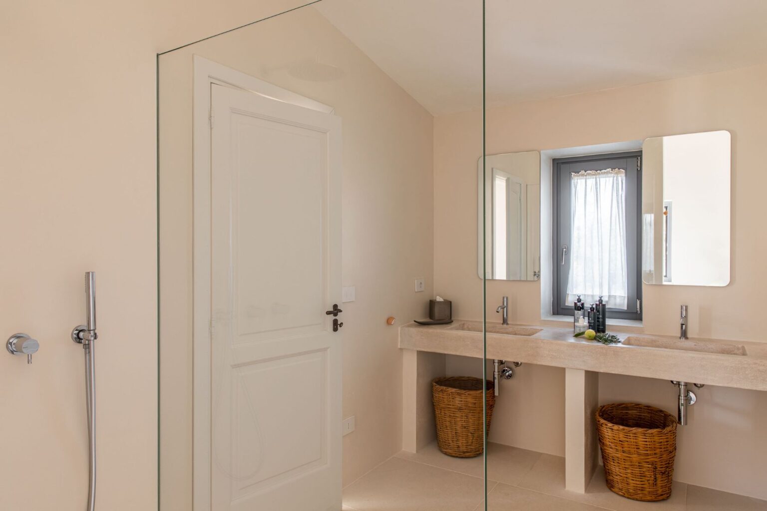 Modern bathroom featuring a double sink made of natural stone, two framed mirrors, and a glass shower enclosure. The neutral-toned walls and floor enhance the spacious feel, while woven baskets beneath the sinks provide stylish storage. A window with sheer curtains allows natural light to brighten the space. | Only on SopranoVillas