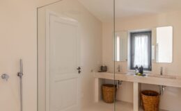 Modern bathroom featuring a double sink made of natural stone, two framed mirrors, and a glass shower enclosure. The neutral-toned walls and floor enhance the spacious feel, while woven baskets beneath the sinks provide stylish storage. A window with sheer curtains allows natural light to brighten the space.