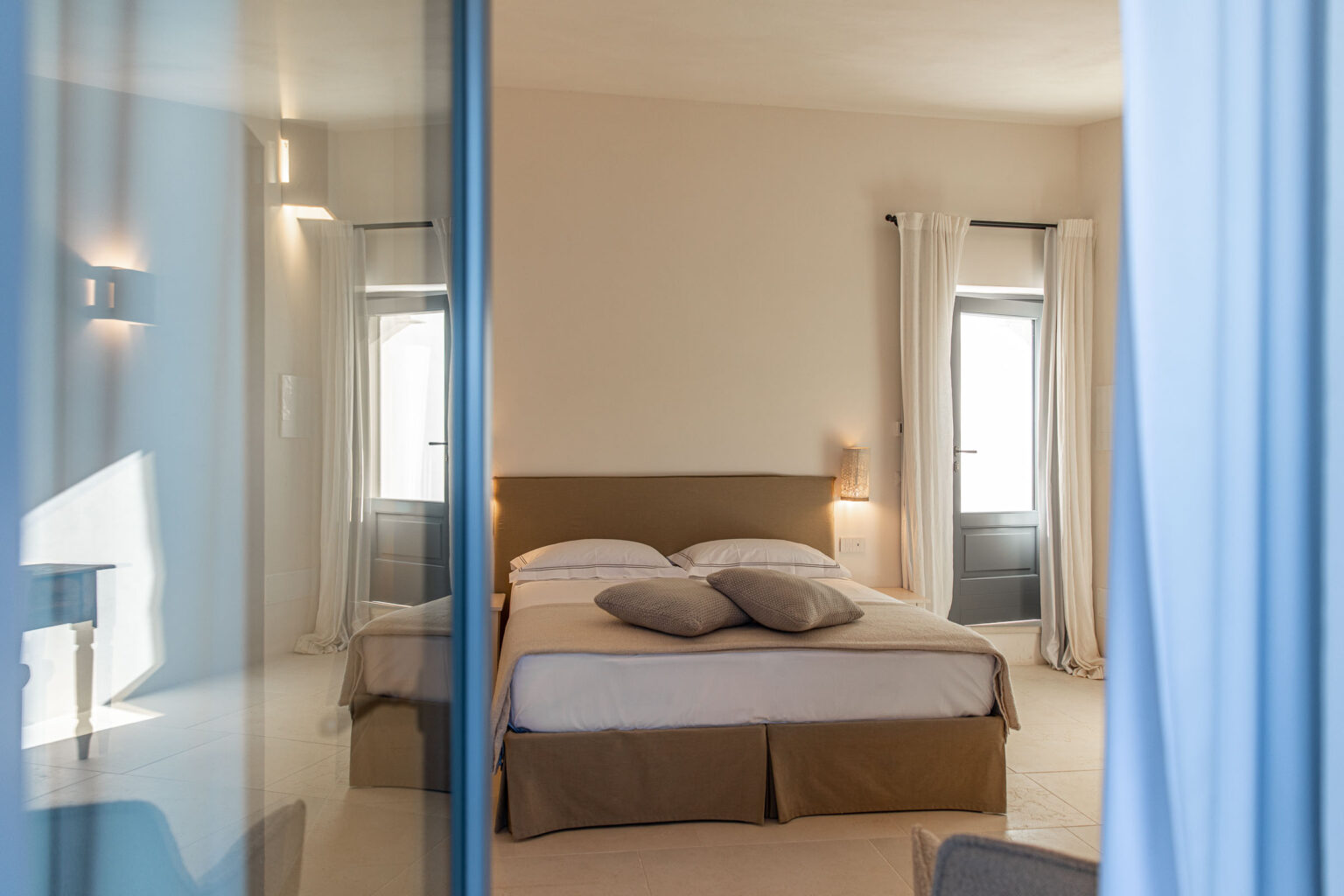 Modern bedroom featuring a cozy double bed with decorative pillows, soft neutral tones, and large windows with sheer curtains. The room is illuminated by stylish wall sconces, creating a serene atmosphere. A glimpse of a blue accent wall and a small wooden table is visible through the glass reflection. | Only on SopranoVillas