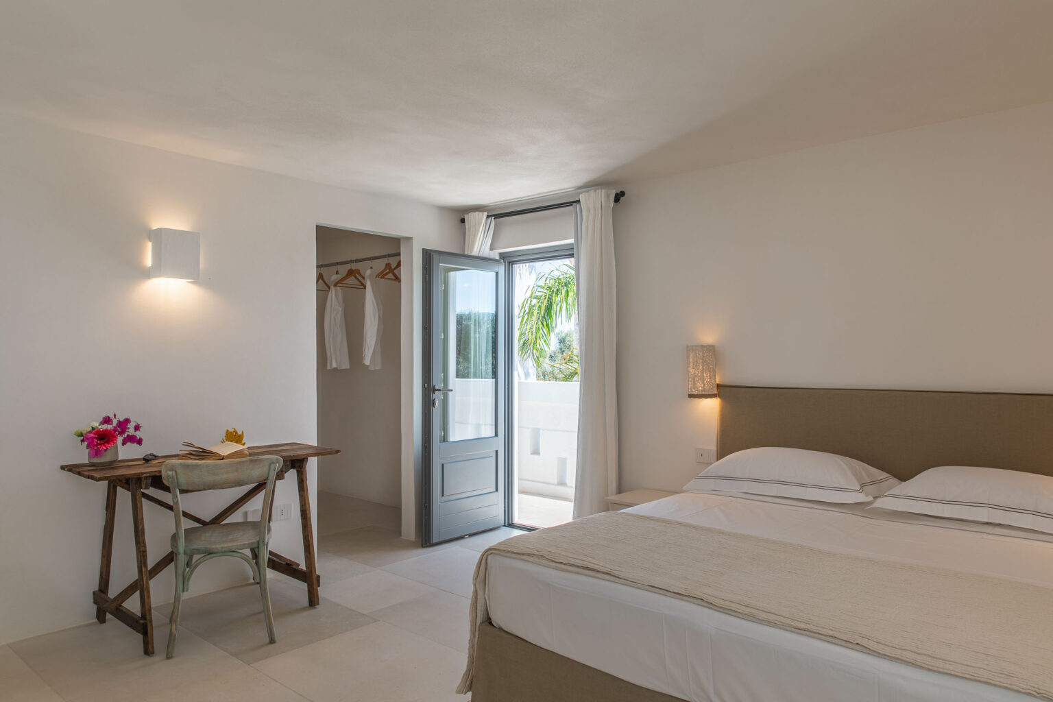 Bright and airy hotel room featuring a comfortable queen bed, a wooden desk with a chair, and a closet with hanging garments. The room includes a door leading to a balcony with views of greenery, complemented by soft lighting and neutral decor. Ideal for relaxation and work. | Only on SopranoVillas