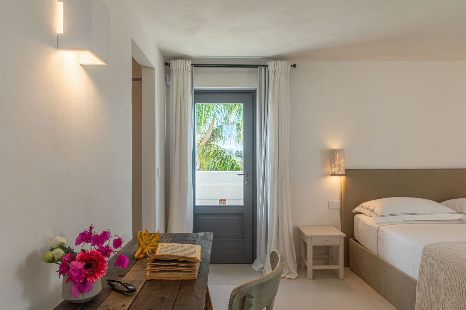 Bright and airy hotel room featuring a cozy bed, wooden desk with flowers and a book, and a door leading to a tropical view. Decor includes soft curtains and modern lighting, creating a relaxing atmosphere. | Only on SopranoVillas