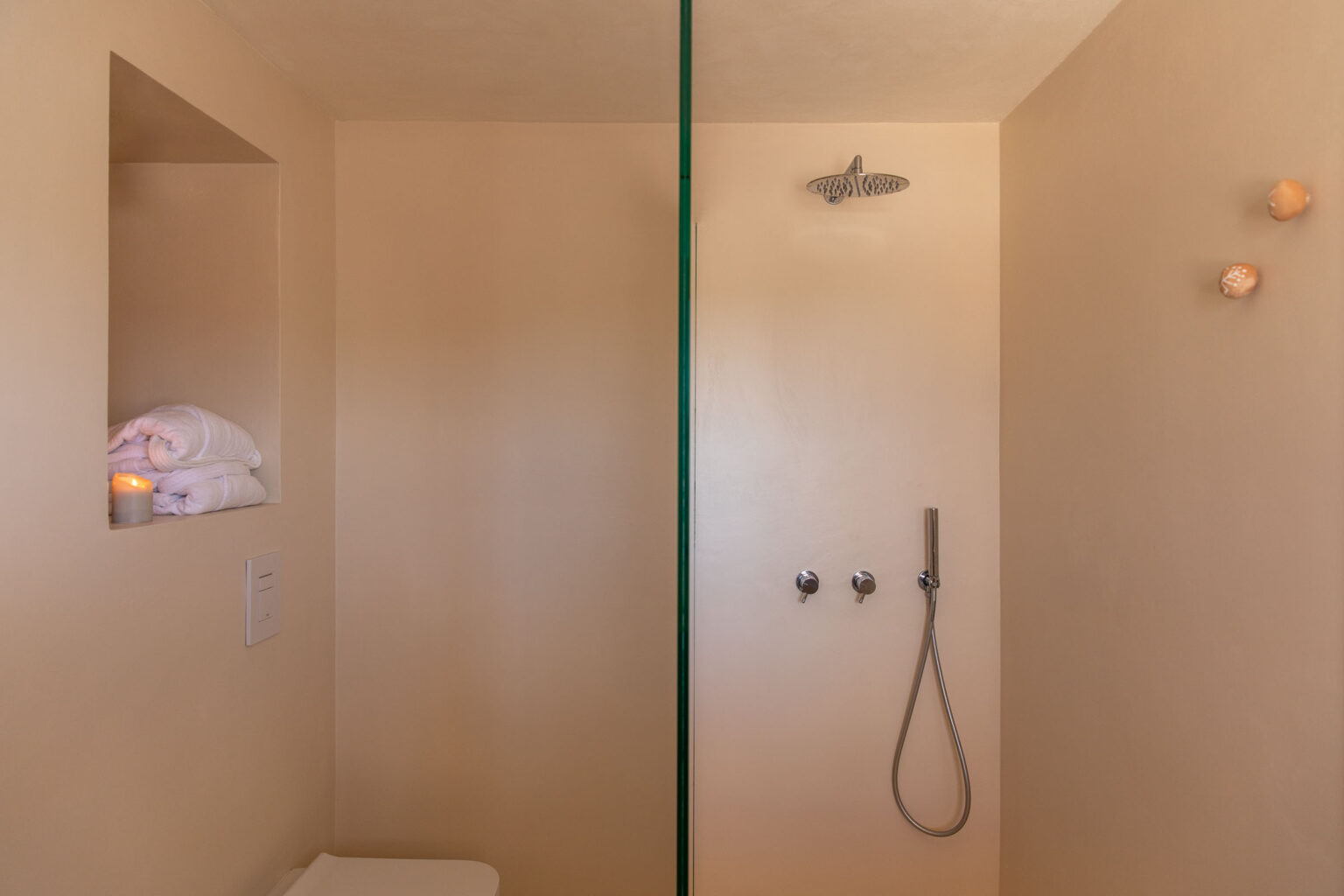 Modern bathroom featuring a spacious shower area with a glass divider, soft peach-colored walls, neatly stacked white towels on a shelf, and a lit candle for ambiance. | Only on SopranoVillas