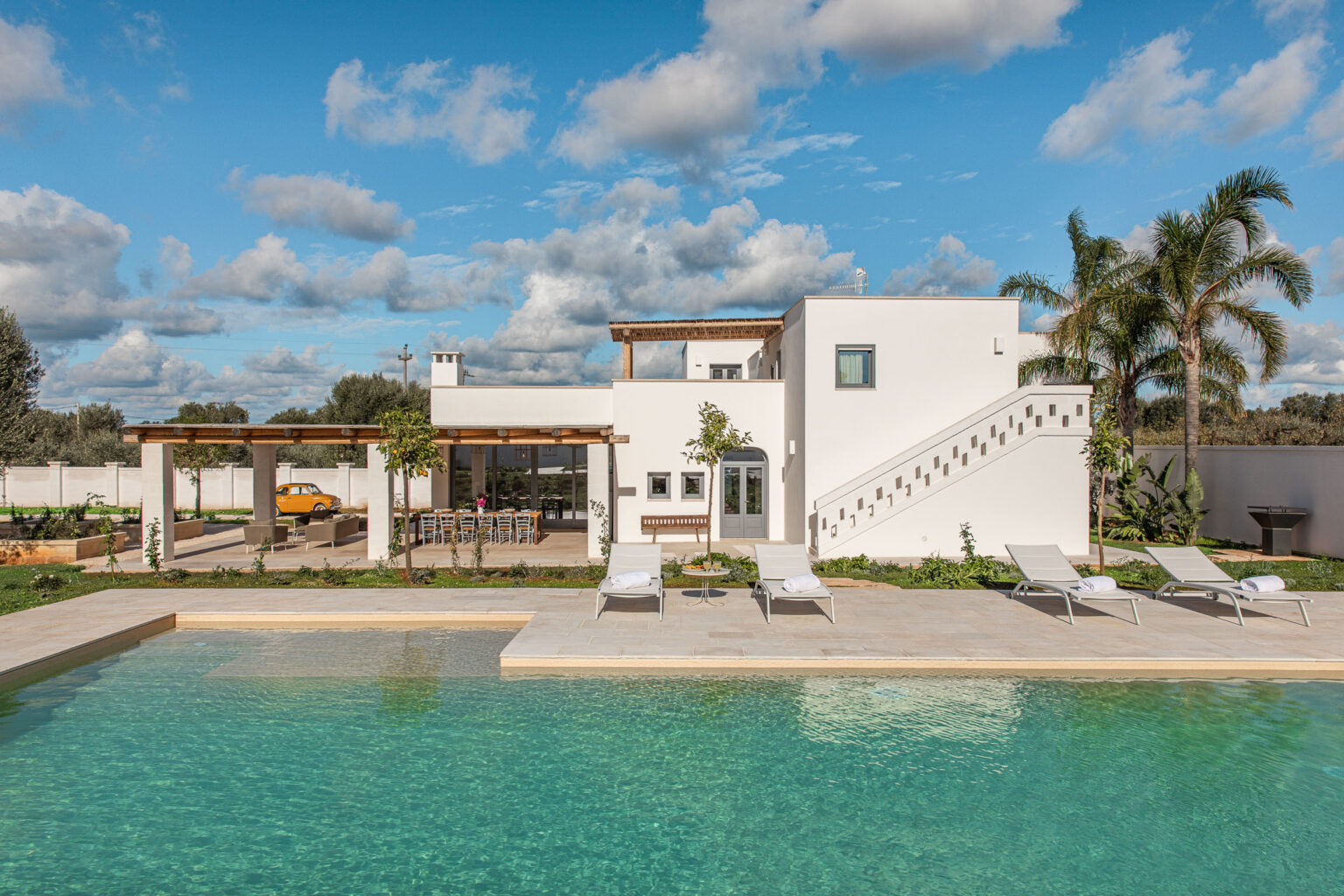 Modern white villa with a pool, surrounded by palm trees and outdoor seating area, under a blue sky with clouds. Ideal for vacation rentals or luxury getaways. | Only on SopranoVillas